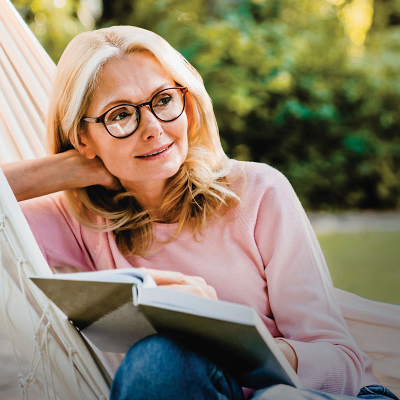 Pre-retired woman sits in a hammock thoughtfully reading a book.